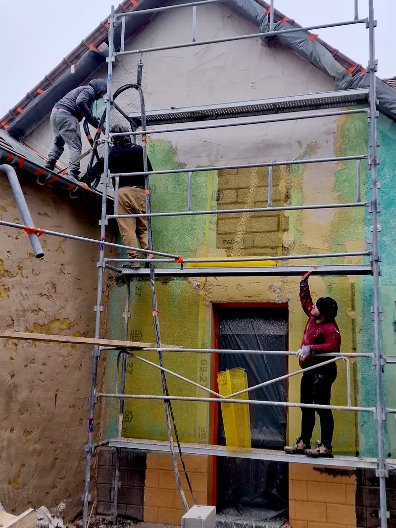 Deux compagnons appliquant le sous-enduit sur le pignon d'une maison de rue, depuis l'échafaudage
