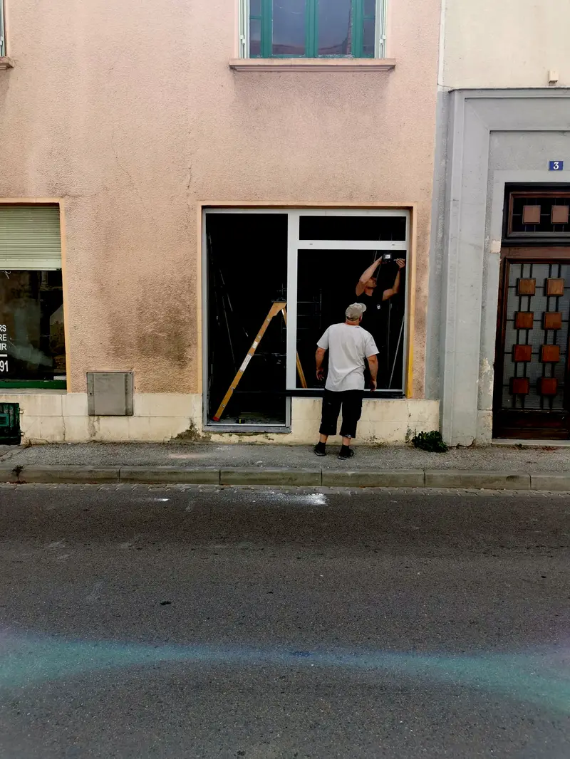 Deux compagnons posent une menuiserie aluminium dans une façade de rue, vue depuis le trottoir d'en face